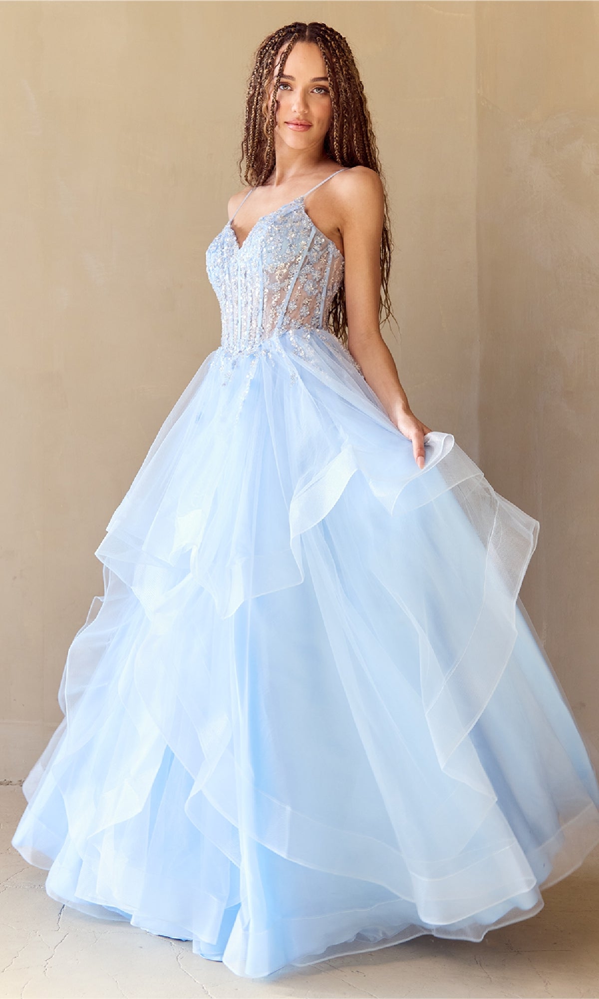 A woman wears the Amelia Couture 5068 Long Glitter Prom Ball Gown, featuring a light blue layered tulle skirt and a fitted beaded bodice with thin straps, standing against a neutral background.